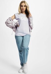 Lyse lilla langærmet top med "DEF" print, kombineret med blå afslappede jeans og hvide sneakers; blødt stof og samlet ærmedetalje.