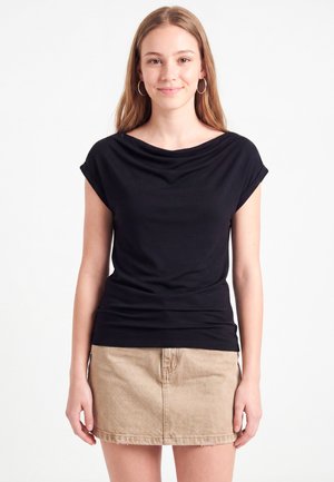 Jeune femme aux longs cheveux châtain clair portant des boucles d’oreilles créoles, un haut noir à manches courtes et une jupe beige, debout sur fond blanc.