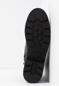Suela de goma negra con textura y diseño de huellas, presenta patrones geométricos elevados y un logo circular en el talón. Perfil lateral liso.