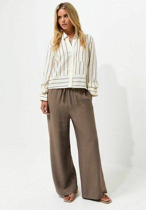 Vrouw met lang blond haar die een witte gestreepte blouse met knopen en losse bruine wijde broek draagt met sandalen.