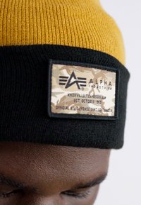 Caciulă tricotată în galben și negru. Are o pată de camuflaj cu text. Material texturat cu o potrivire strânsă.