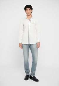 Vit långärmad skjorta med svart kant, liten logotyp på bröstet, i kombination med ljusblå jeans och svarta skor. Enkel design och åtsittande form.