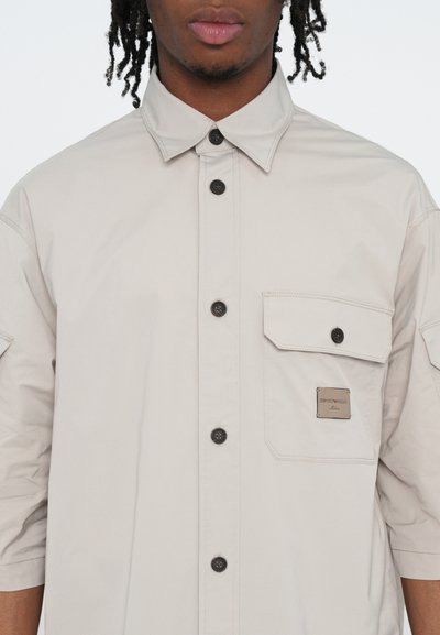 Chemise beige clair à manches courtes avec boutonnière sur le devant, poche poitrine avec étiquette, et boutons noirs en contraste. Texture de tissu lisse.