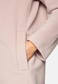 Helles rosa Mäntelchen aus weichem Stoff, mit einer Seitentasche und abgerundeten Ärmeln. Glatte Textur mit minimaler Detailsgestaltung, keine sichtbaren Verschlüsse.