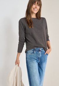 Maglietta a maniche lunghe a righe grigio scuro con vestibilità rilassata, abbinata a jeans in denim blu chiaro. Un maglione di colore crema con texture è tenuto in una mano.