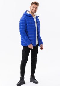 Giacca a vento blu con cappuccio, caratterizzata da cuciture orizzontali, una tasca con zip e polsini elastici. Indossata sopra un felpa bianca e pantaloni cargo neri.