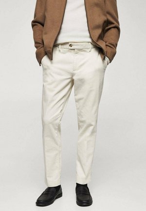 Mann trägt beige Cordhosen, braune Jacke, weißes Hemd und schwarze Loafers, steht mit Händen in den Taschen.