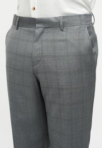 Gros plan sur un pantalon gris sur mesure avec passants de ceinture, porté avec une chemise blanche rentrée montrant un bouton marron visible.