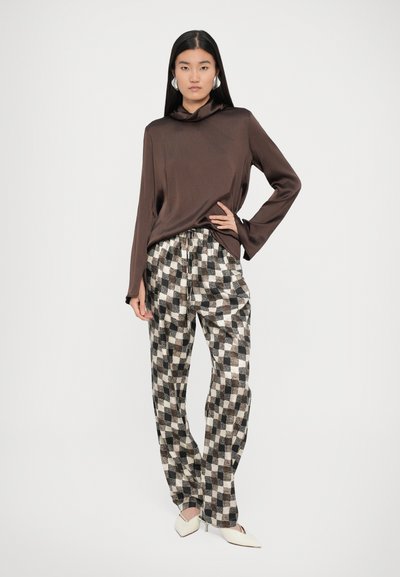 Top lung cu mâneci lungi din satin maro, cu guler drapat, asortat cu pantaloni în carouri alb-negru. Modelul poartă pantofi cu vârf ascuțit de culoare crem.