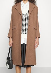 Manteau en laine marron avec un col cranté, deux boutons à l'avant et des poches plaquées. Pull en pied-de-poule noir et blanc porté en dessous.
