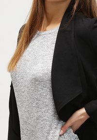 Blazer leggero nero con fronte drappeggiato sopra un top in maglia grigia. Tessuto testurizzato, forma snella e design a frono aperto.