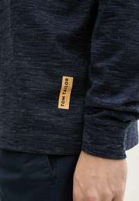 Námornícky textúrovaný dlhý rukáv tričko s malou béžovou nášivkou s nápisom "TOM TAILOR" na ľavej strane. Jednoduchý, minimalistický dizajn.