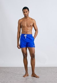 Μπλε μαγιό σορτς με λευκό κορδόνι, κατασκευασμένο από ελαφρύ ύφασμα. Διαθέτει μικρό λογότυπο στο αριστερό πόδι. Το μοντέλο στέκεται σε υφή δαπέδου.