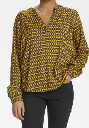 Frau trägt eine gelb-braune, diamantgemusterte Langarmbluse mit V-Ausschnitt, kombiniert mit schwarzen Hosen, eine Hand in der Tasche.