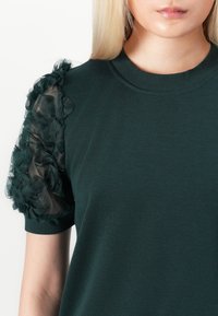 Mørkegrønn topp med korte ermene pyntet med lagdelte svarte tylblomster. Glatt tekstur med ribbet rund halslinning.