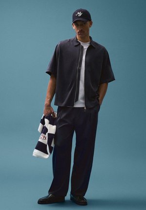 Uomo con cappellino nero, maglia a maniche corte nera con zip, t-shirt bianca, pantaloni neri, che tiene un maglione a righe su sfondo verde acqua.