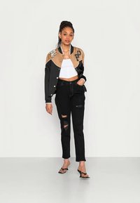 Jaqueta varsity preta curta com acentos em tons de bege, top curto branco, jeans pretos desgastados e sandálias pretas. Tecido suave com texturas contrastantes.