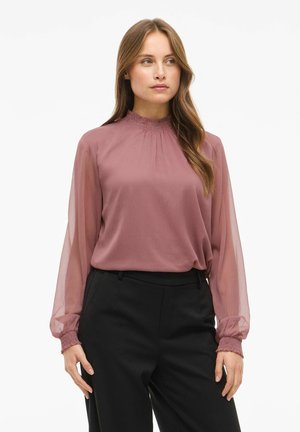Blusa de manga larga en malva con un escote fruncido y mangas transparentes. La tela es ligera con una ligera textura. Combinada con pantalones negros.