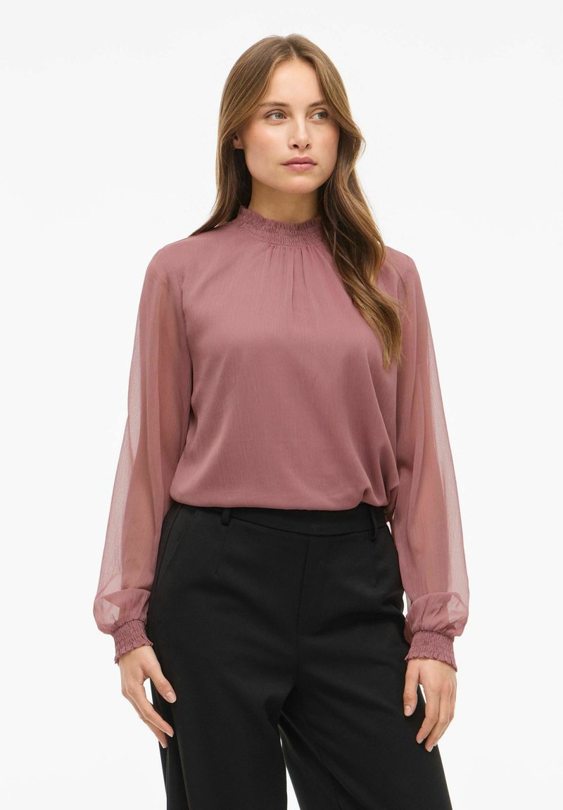 Blusa de manga larga en malva con un escote fruncido y mangas transparentes. La tela es ligera con una ligera textura. Combinada con pantalones negros.