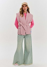 Prošívaná růžová vesta s páskem a předními kapsami, přes jasně růžový svetr. V kombinaci se světle modrými širokými džínami a teniskami.