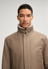 Braune Jacke mit hohem Kragen, mit einem weichen, hellen Futter ausgestattet, Druckknopfverschluss und Seitentaschen; glatte Stofftextur.