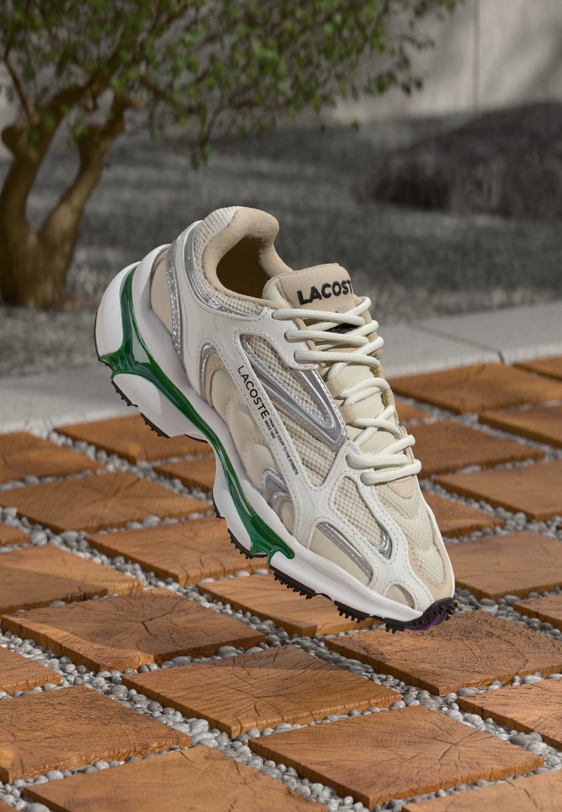 Beige och vit Lacoste-sneaker med gröna sula accenter, sväva över en träplatta och grusig mark utomhus nära ett litet träd.
