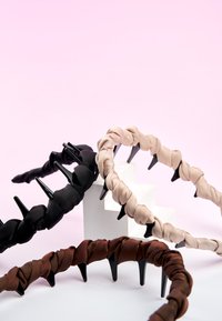 Trois serre-têtes en plastique enveloppés de tissu beige, noir et marron disposés sur des blocs géométriques blancs sur fond rose.