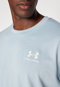 Světle modré bavlněné tričko s kulatým výstřihem a bílým logem Under Armour na hrudi. Hladký povrch, ležérní střih.