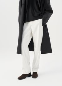 Cappotto nero chiuso con bottoni sopra una camicia nera, abbinato a pantaloni bianchi e scarpe in suede marroni. Il cappotto ha una vestibilità rilassata e una texture morbida.