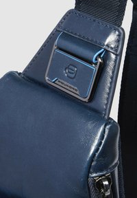 Borsa a tracolla in pelle blu navy con texture liscia, ferri metallici, dettagli blu a contrasto e tracolla regolabile. Presenta il logo del marchio.