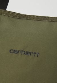 Sac en tissu vert olive avec des coutures noires et un logo "carhartt" brodé. Texture lisse sans motifs visibles ni matériel supplémentaire.