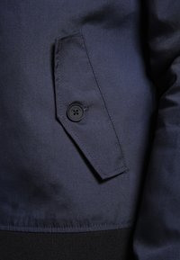 Námořnická modrá bunda s klopovou kapsou a černým tlačítkem, hladká textura a elastický černý pas.