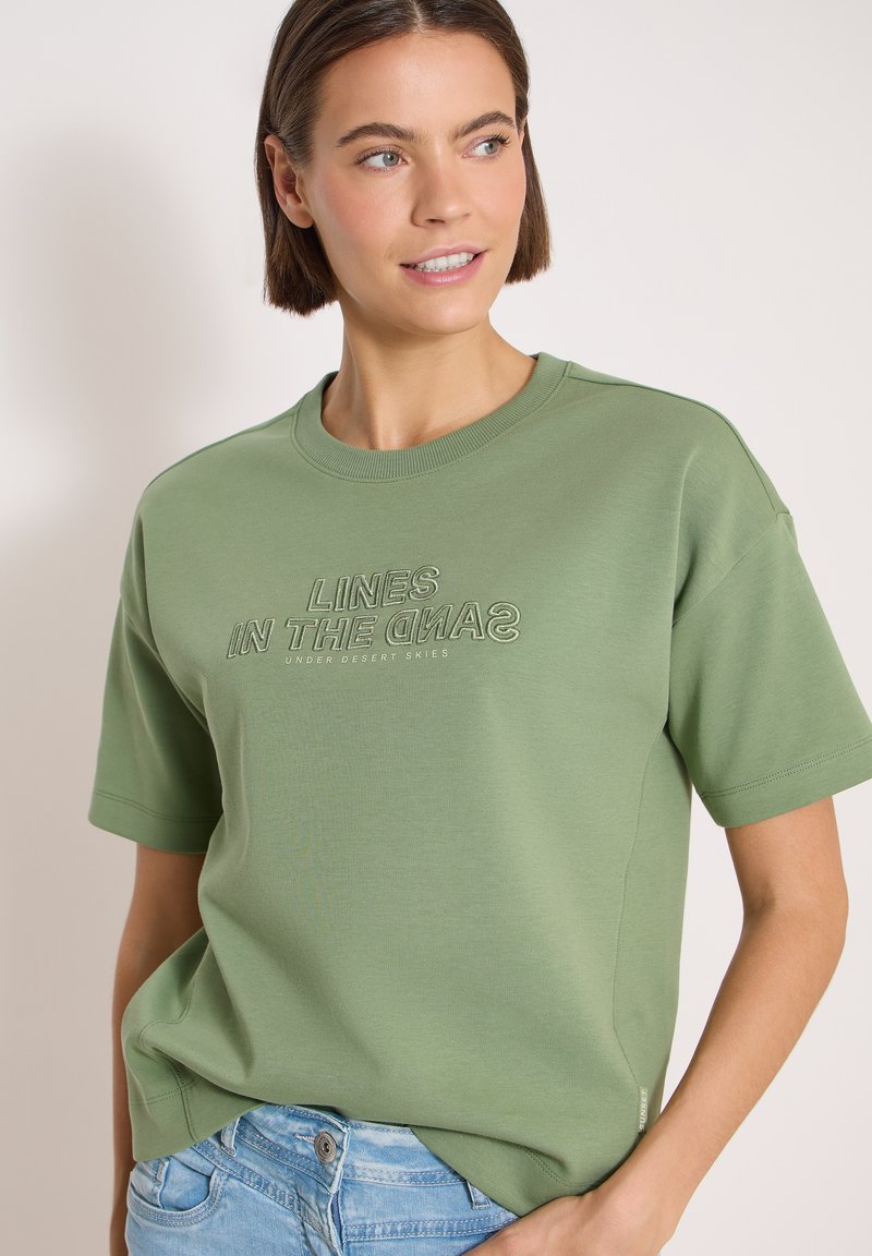 Femme portant un T-shirt vert olive avec le texte "LINES IN THE SAND UNDER DESERT SKIES", associé à un jean bleu clair, regardant sur le côté.