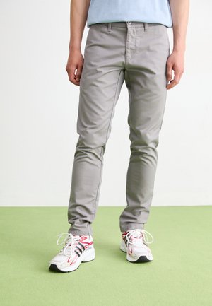 Mann trägt hellgraue Slim-Fit-Hose und weiße Sneakers mit roten und schwarzen Akzenten, steht auf einer grünen Fläche.
