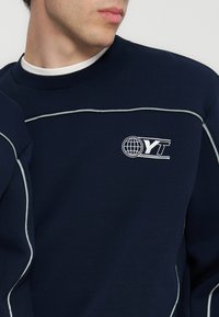 Άντρας που φοράει μπλε φούτερ με λευκή επένδυση και λογότυπο με σφαίρα και τα γράμματα "YT" στο στήθος.