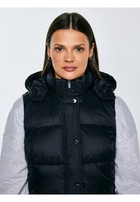 Sirgete tumedate juustega naine kannab helehallil dressipluusil musta sulepalli vestit ja vaatab otse ette neutraalse ilmega.