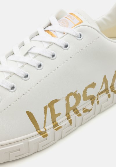 White leather sneaker featuring a gold "VERSACE" print, round toe, and flat white laces with a textured, patterned rubber sole.