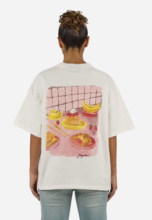 Femeie purtând un tricou alb cu o ilustrație colorată a unor mâncăruri de mic dejun, inclusiv clătite, banane, croissante, căpșuni și o ceașcă de cafea.