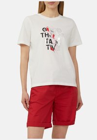 Witte katoenen t-shirt met korte mouwen met zwarte en rode tekst: "GOEDE DINGEN HEBBEN TIJD NODIG"; gecombineerd met rode shorts.