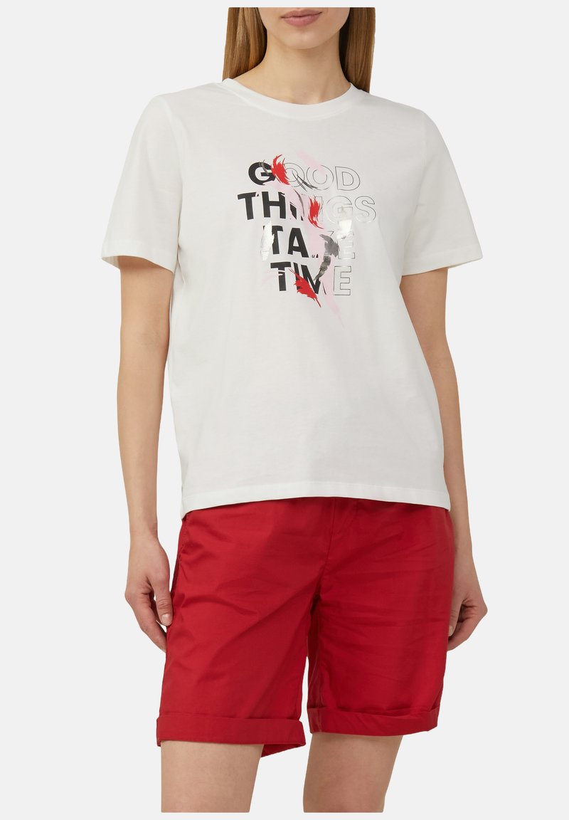 Witte katoenen t-shirt met korte mouwen met zwarte en rode tekst: "GOEDE DINGEN HEBBEN TIJD NODIG"; gecombineerd met rode shorts.