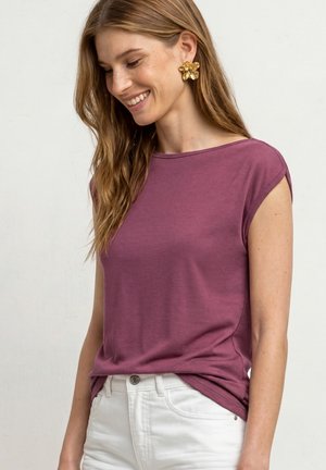 Femme souriante, portant un haut violet à manches courtes, un pantalon blanc et de grandes boucles d'oreilles dorées en forme de fleur, debout devant un fond clair uni.