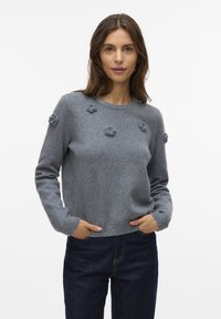 Frau trägt einen grauen Pullover mit kleinen gestrickten Blumenverzierungen und dunklen blauen Jeans, steht mit den Händen in den Taschen vor einem schlichten weißen Hintergrund.