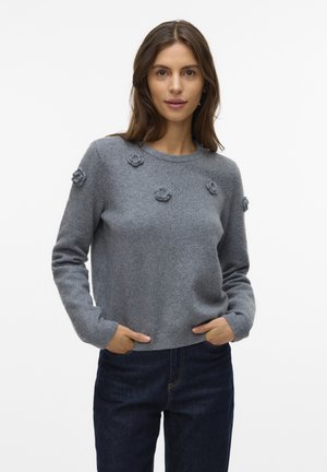 Mujer vistiendo un suéter gris con pequeños detalles de flores de punto, jeans azul oscuro, de pie con las manos en los bolsillos contra un fondo blanco liso.