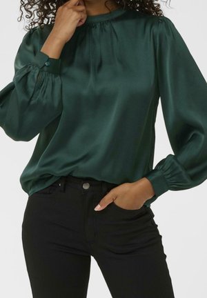 Femme portant un chemisier en satin vert foncé avec de longues manches bouffantes et un pantalon noir, une main près du visage et l'autre dans la poche.