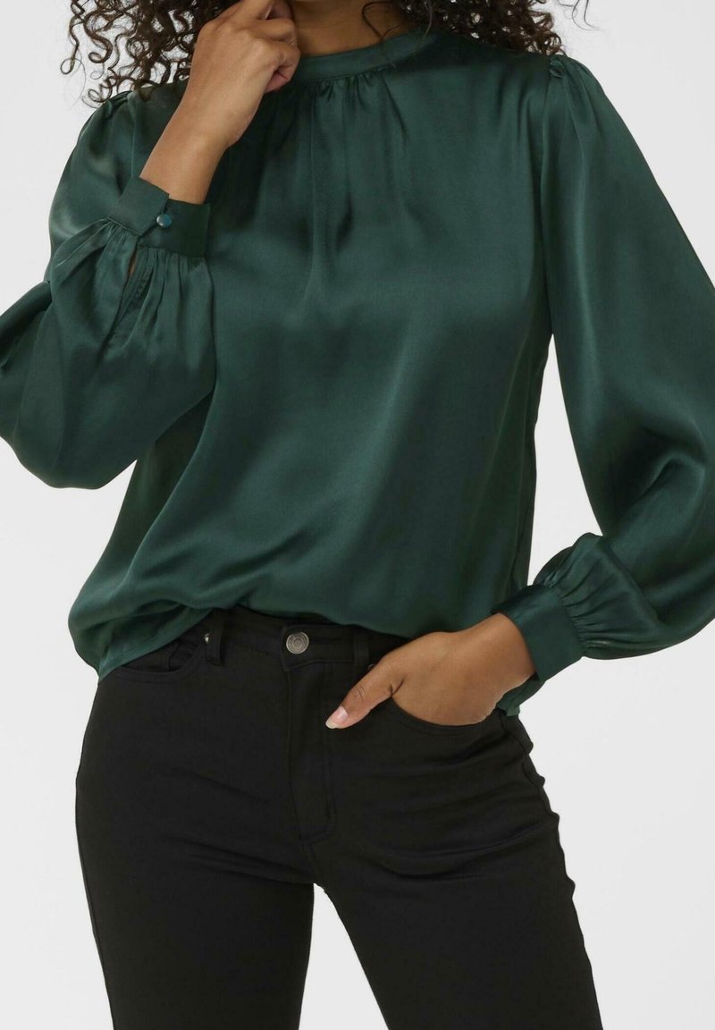 Femme portant un chemisier en satin vert foncé avec de longues manches bouffantes et un pantalon noir, une main près du visage et l'autre dans la poche.
