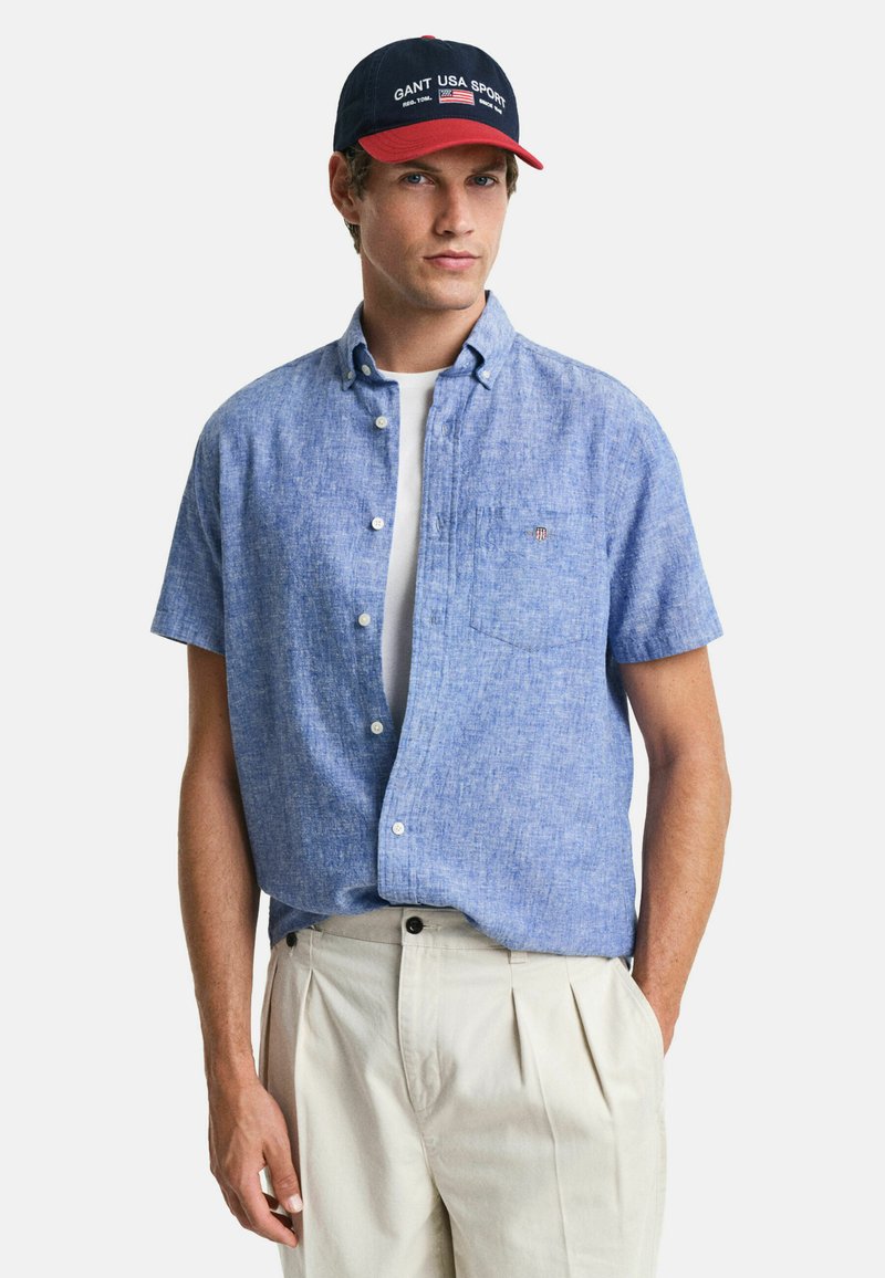 Korte mouwen blauwe linnen shirt met knoopsluiting, linker borstzak, gecombineerd met lichtbeige geplooide broek en een marineblauwe/rode pet.