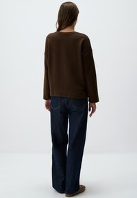 Maglione brown oversize con scollatura rotonda abbinato a jeans a gamba larga in denim scuro e scarpe beige senza lacci, visto da dietro.
