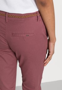 Dopasowane bordowe spodnie z plecionym brązowym paskiem. Posiadają tylną kieszeń z metką logo oraz gładką teksturę materiału.