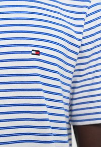 Λευκό πουκάμισο με λεπτές οριζόντιες μπλε ρίγες και μικρό ορθογώνιο κόκκινο, λευκό και μαύρο κέντημα λογότυπου στο στήθος.