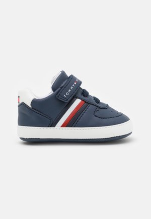 Zapatilla de niño azul marino con suela blanca, rayas laterales rojas y blancas, y una correa de velcro con la etiqueta "Tommy Hilfiger".